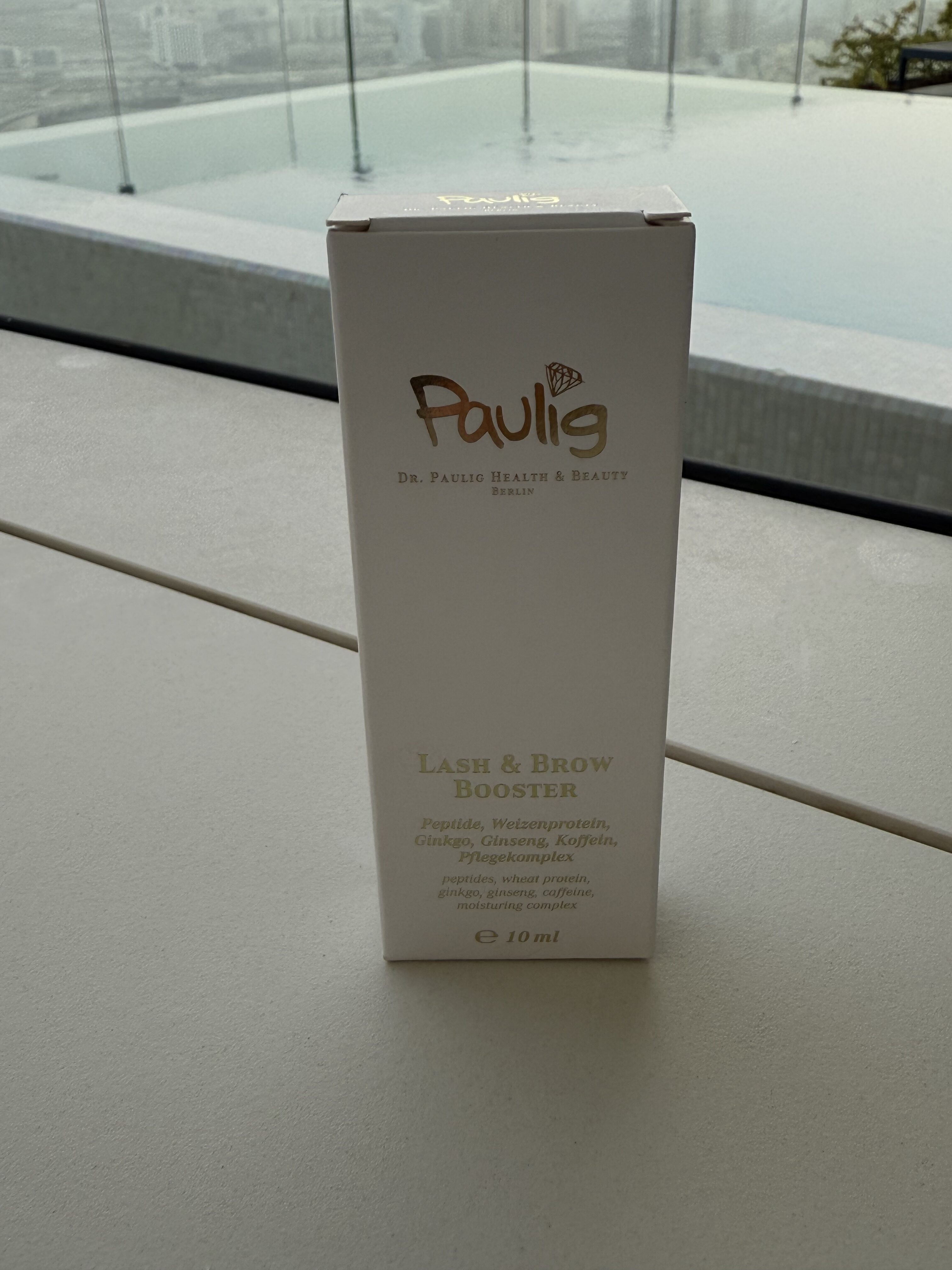 Eine kleine, weiße Schachtel mit der Aufschrift "Lash & Brow Booster" steht auf einem Tisch vor einem Pool mit Stadtblick im Hintergrund.